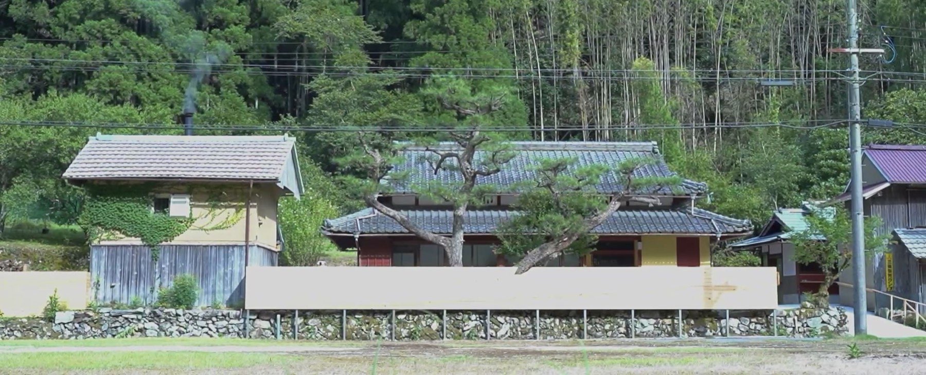 田んぼの向こうに建つ母屋と蔵。蔵の煙突から白い煙が上がる