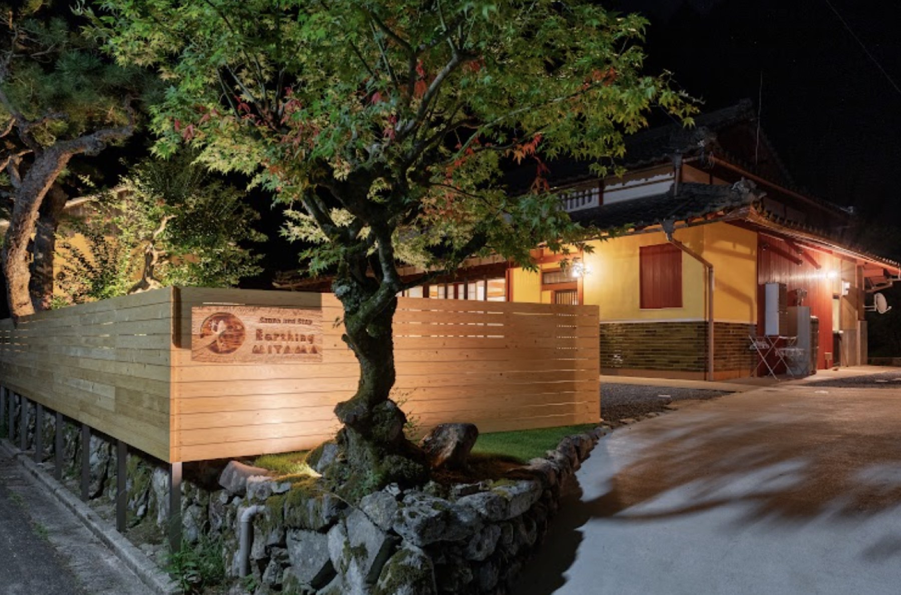 夜。木の看板と松が照らされ、母屋から灯りが漏れる
