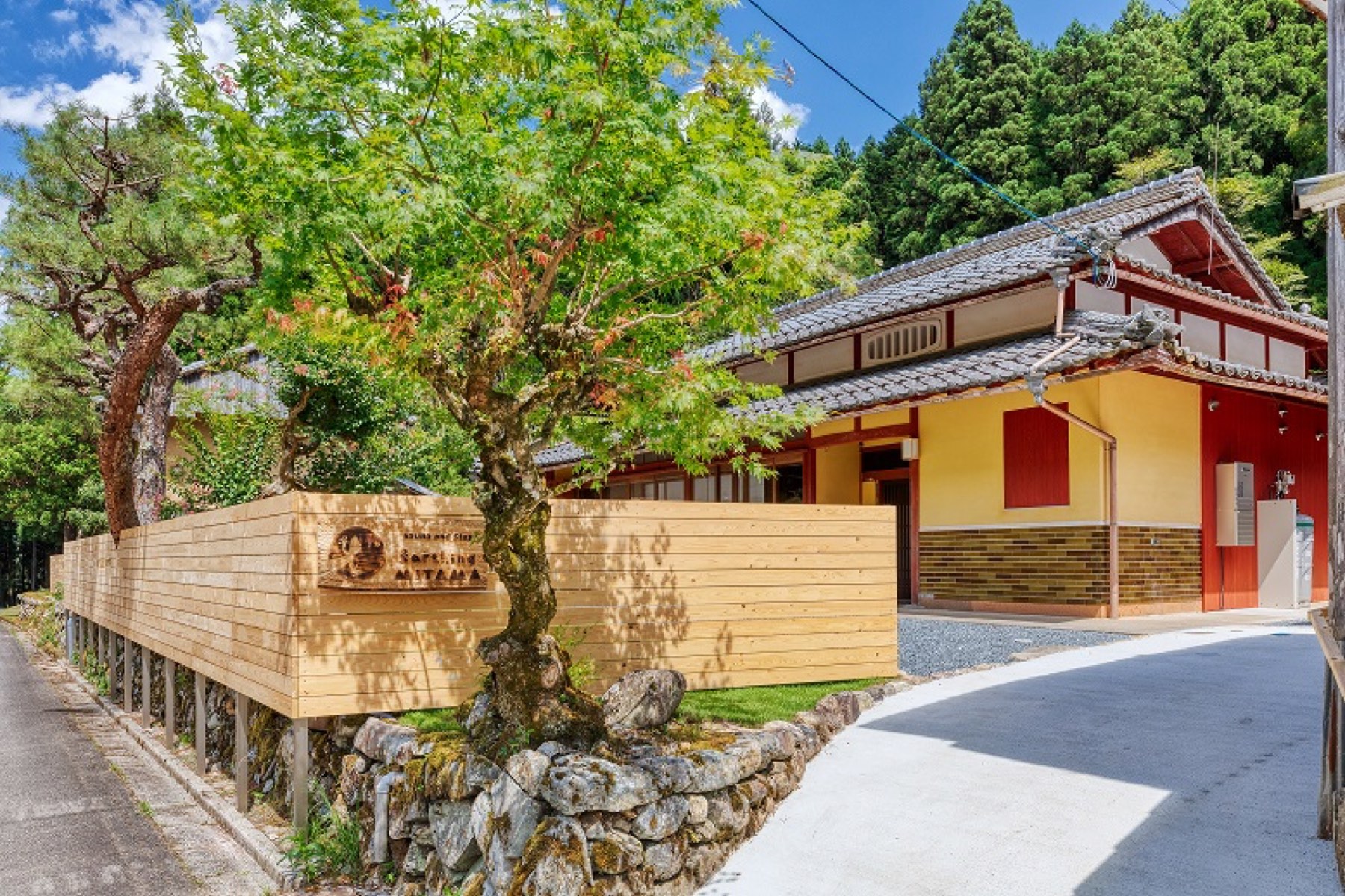 弁柄色の受付棟と芝生の庭。手前に松の木と木の看板、奥に山