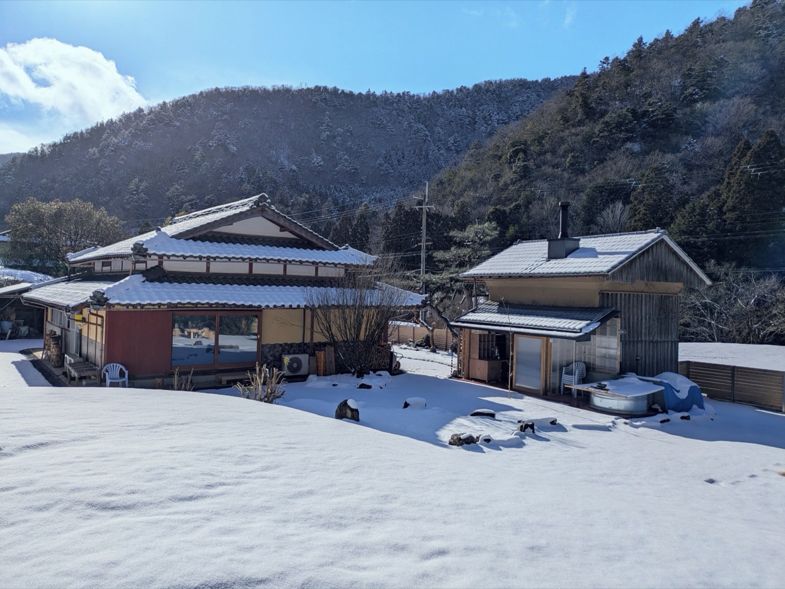 雪の積もった母屋と蔵。屋外の水風呂にも雪が載り、背後の山も白い